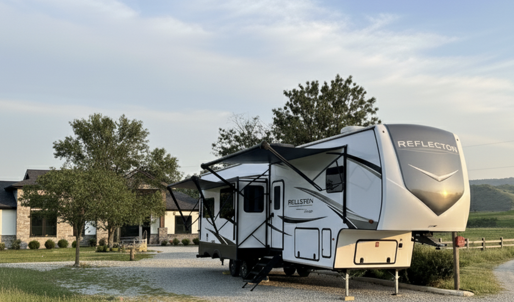 Fifth-wheel RV parked on a rural gravel pad with rolling green hills and a farmhouse in the background.